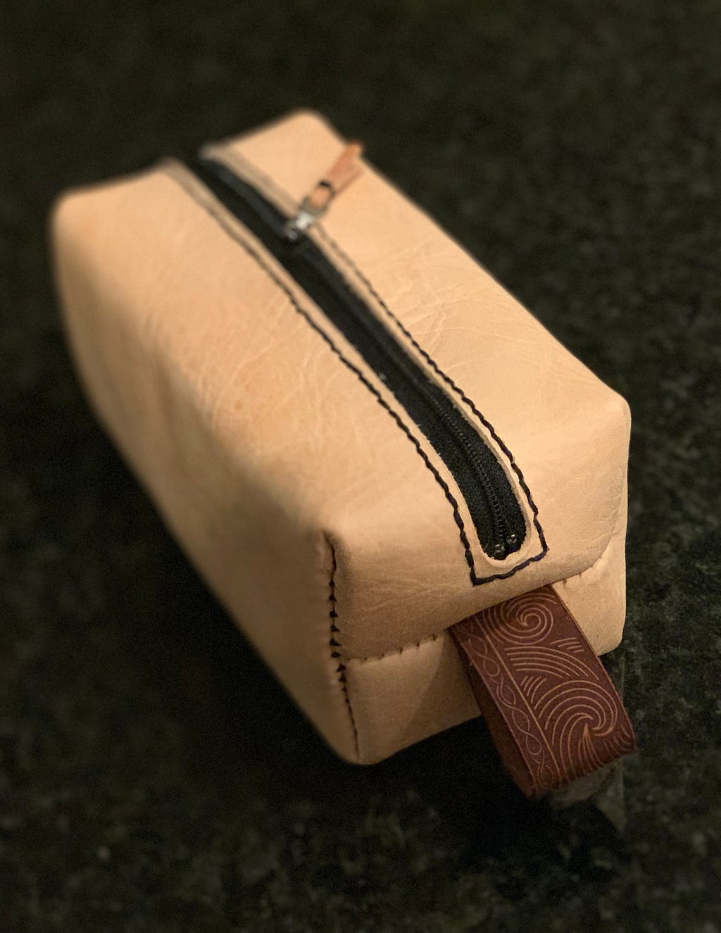 A tan colored leather carrying case, a soft rectangle about the size of a luggage toiletry carrier. There’s a black zipper down the middle, and a leather loop on the side with an intricate, almost ancient pattern. (In all honesty it’s the pattern on Iron Fist Alexander, Warrior Jar in the game Elden Ring.)