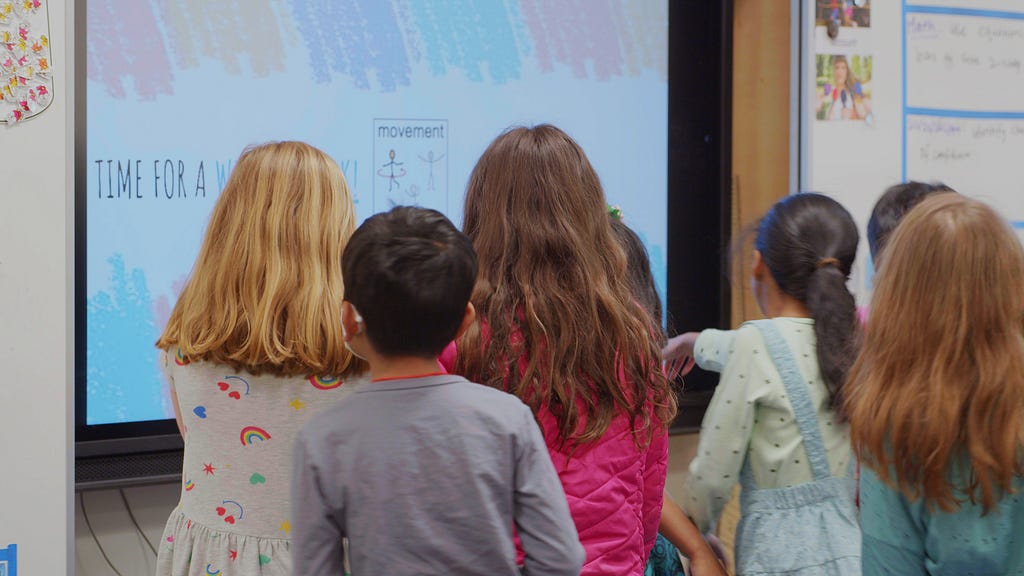 A group of elementary school children, with their backs facing the viewer, work on a large screen in their classroom.