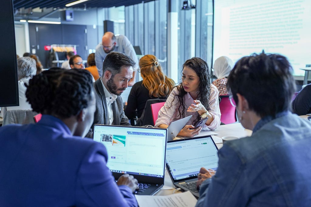 Four people sit at a desk with laptops. One person is talking.