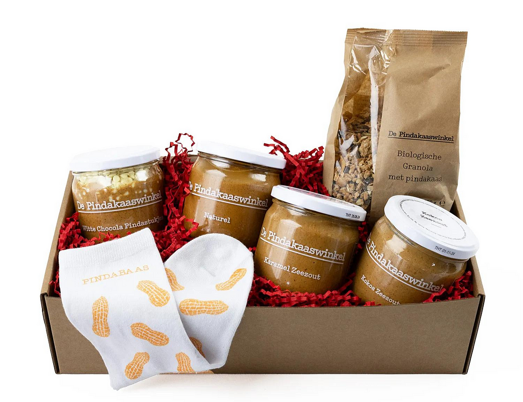 A cozy holiday-themed box from ‘De Pindakaaswinkel’ packed with jars of peanut butter, ready for gifting. The box, with a see-through window, showcases the jars ready to be shared. The picture captures the holiday season, where simple delights like peanut butter become gifts of joy, wrapped in the warmth of the season.