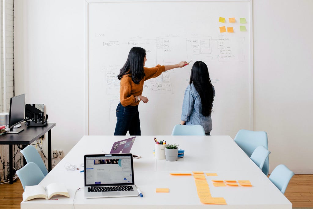 An image of two women standing in from of a whiteboard with stick-it notes pasted on the board. One of the women is pointing to the board and explaining something to the other woman. The women are in an office setting, there is a laptop, two mugs and some sticky notes on the table.