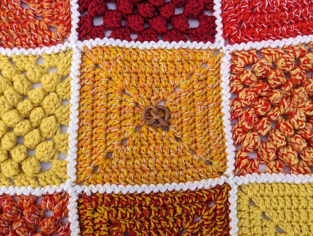 Square is made out of yellow and orange yarn with white embroidery floss. A light brown clay button in the shape of a pretzel is stitched into the centre of the square.