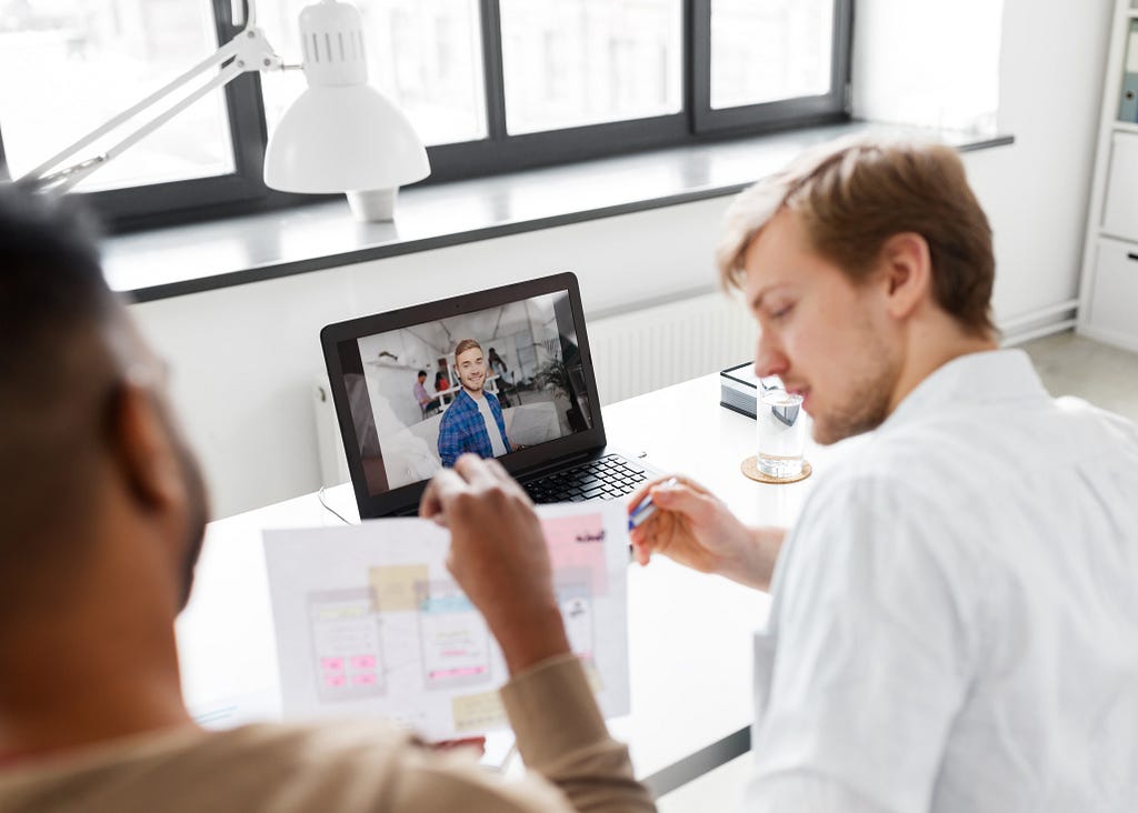 Two designers reviewing a paper prototype with sticky notes while conducting a remote customer interview with a participant visible on the laptop screen