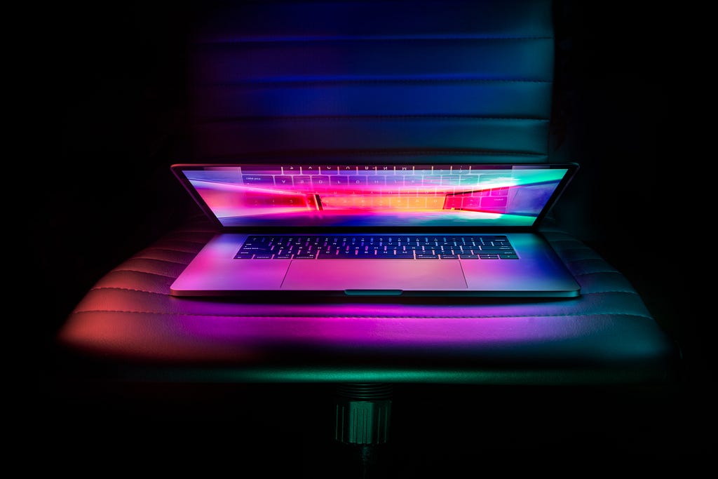 A partially open Apple laptop with bright colors pouring out sits on a chair in a dark room, courtesy Ash Edmonds Koxa, Unsplash (GX_5zs)