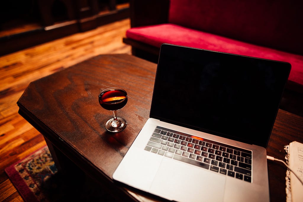 This visual depicts a woody living room table with a laptop on it, besides is a glass full of wine.