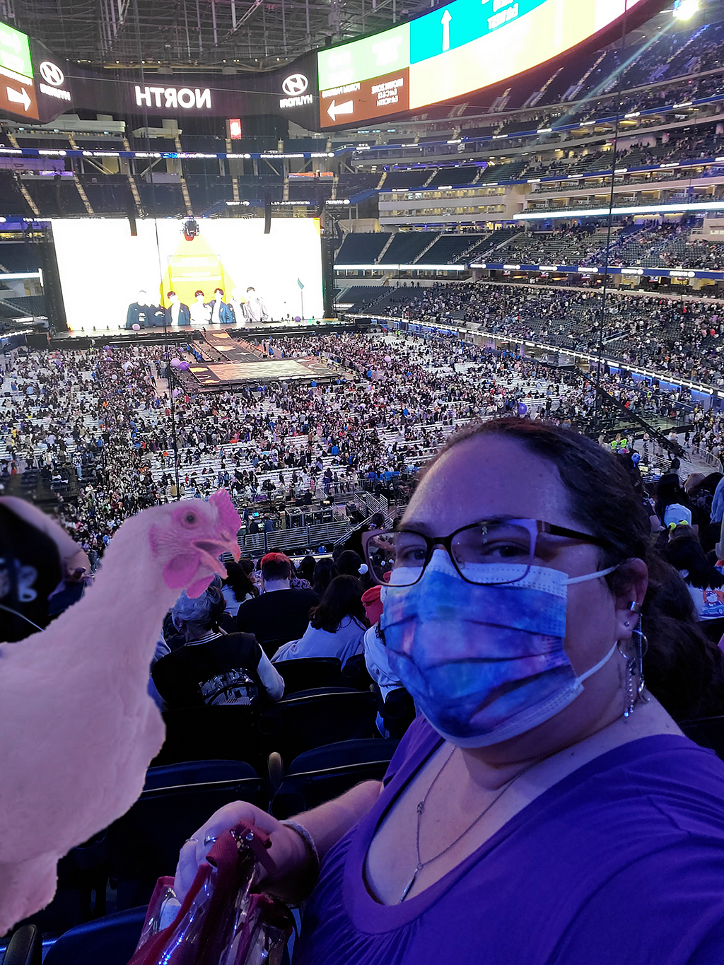 Selfie at a stadium with a woman with a purple face mask and purple shirt on next to a purple colored chicken badly photoshopped in
