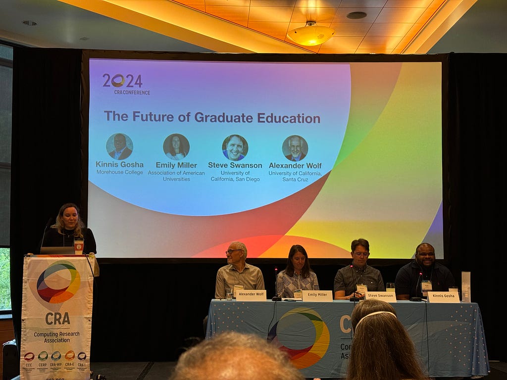 A podium with Gillian, a table with Kinnis, Emily, Steve, and Alexander, and a big screen with the phrase “The Future of Graduate Education”