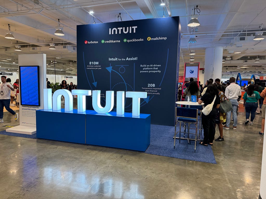 Conference attendees standing in line to play basketball in a large vendor space with an oversized light-up “Intuit” branded sign.