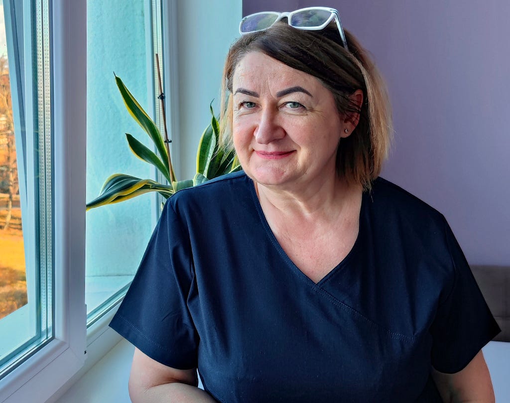 A woman in navy surgical scrubs leans against a window in a hospital room.