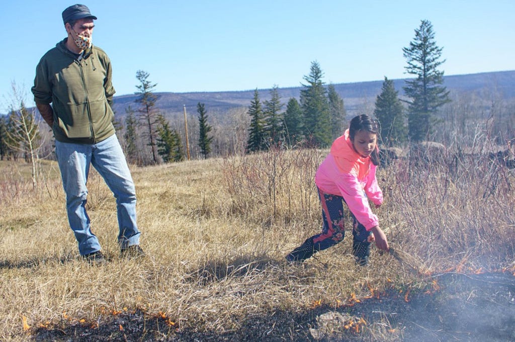 A man watches while his daughter adds some long grass to a controlled burn on the ground.