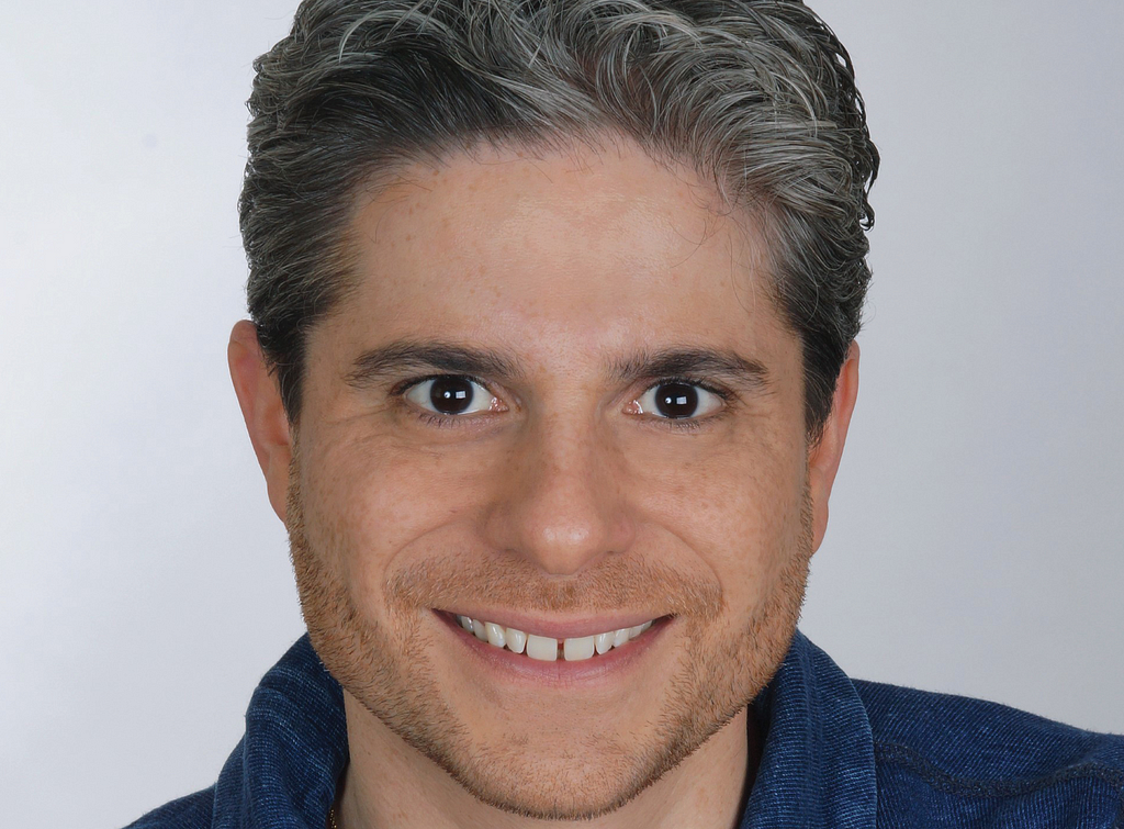 A man with salt and pepper hair and a blue shirt smiles for the camera