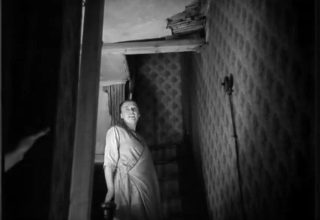 A still from Housing Problems (1935) .A woman stands at the top of crooked stairs in her house. A hole in the ceiling is visible above.