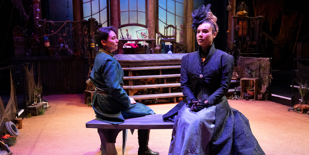 Two performers sit on a bench on a stage, one in a teal jacket and one in 19th century-inspired mourning dress.