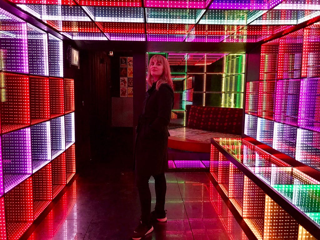 A woman stands in a brightly lit art exhibit-style room. There are colorful cubes emitting red, purple, white, green, and orange lights.