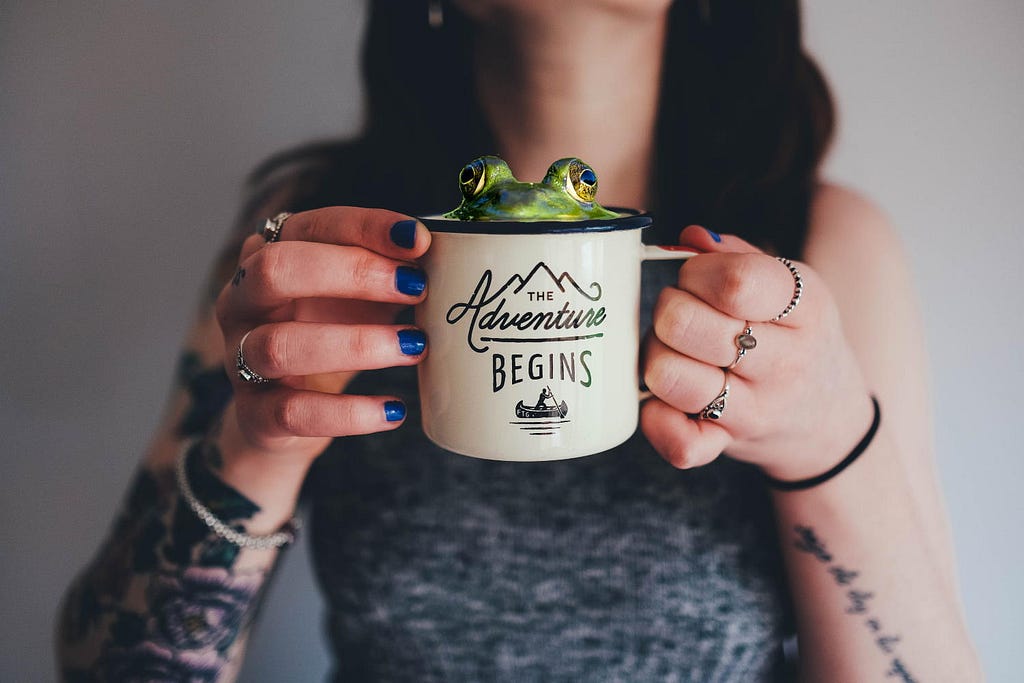 A tattooed woman holds a coffee mug with “The Adventure Begins” written on it. A frog is peeking out of the top of the mug.