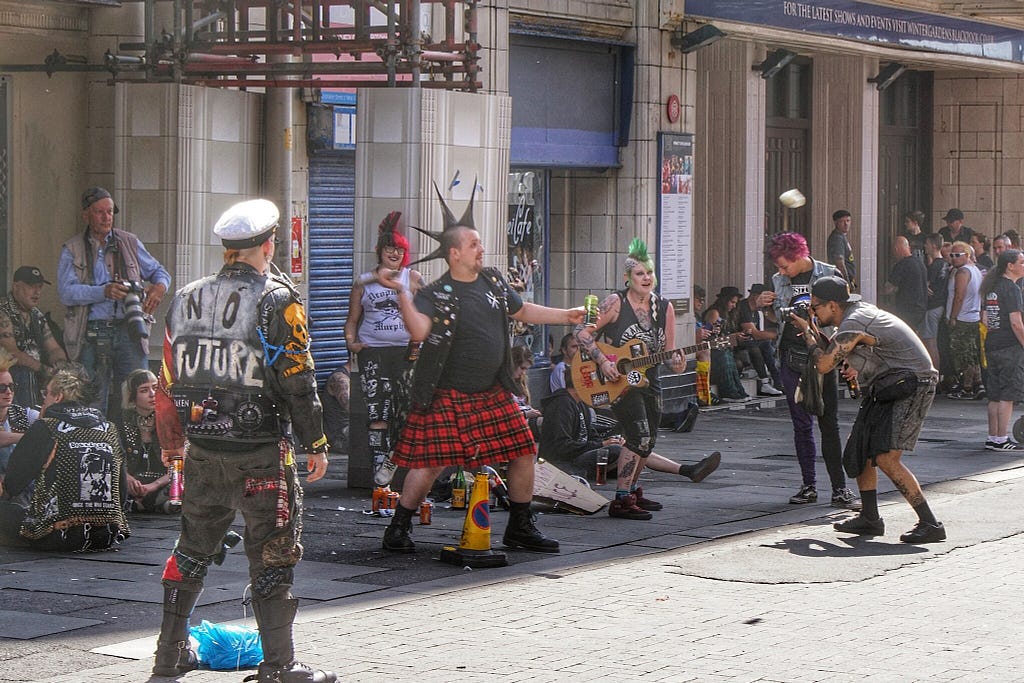 Punks on the street. Some are playing music, one is holding a bottle and a baseball bat while another one is taking a picture. There are “normal” photographers and bystanders in the background.