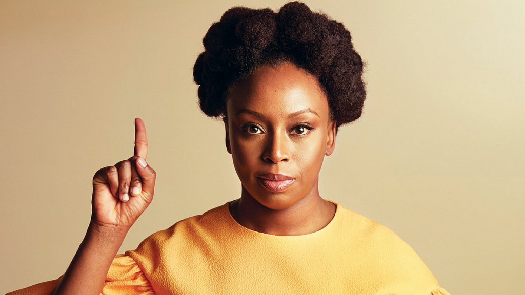 Imagem da escritora Chimamanda Ngozi Adichie de vestido amarelo, com o dedo indicador levantado e olhando para a câmera.