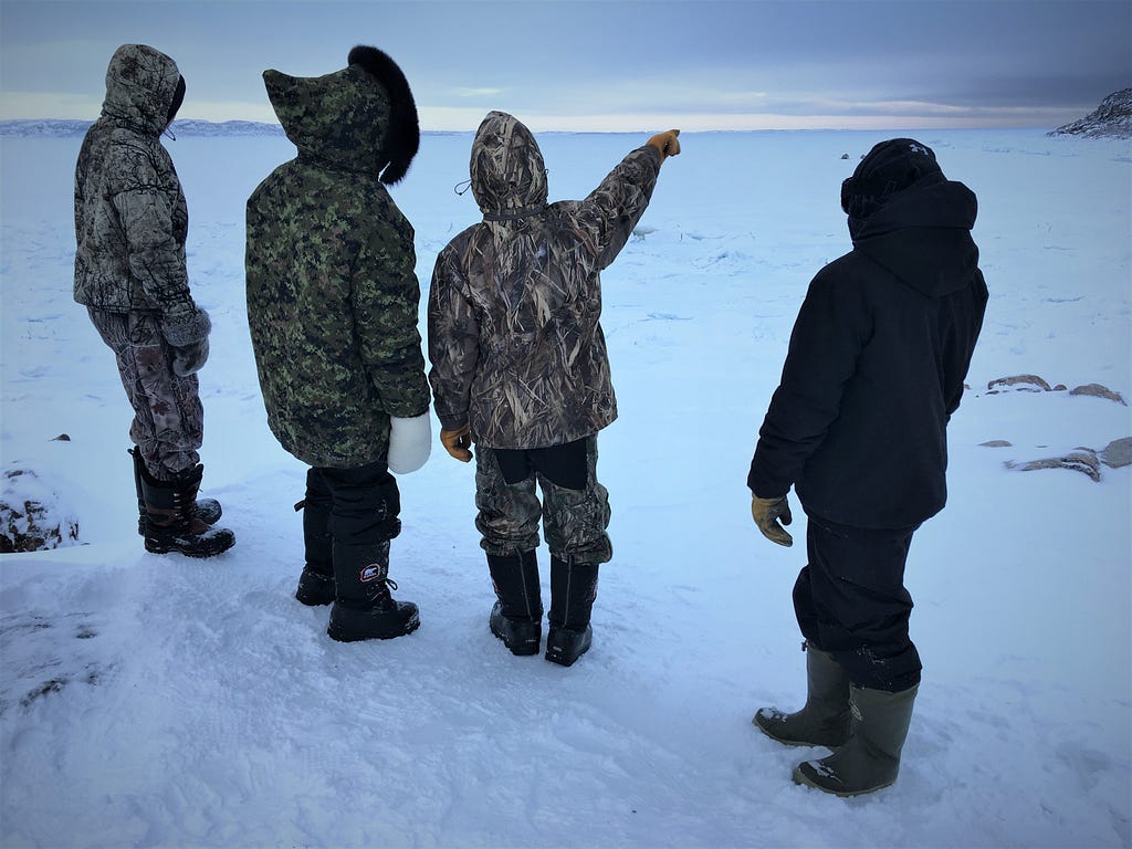 Four people dressed in winter clothing on snow. One person is pointing at the horizon.