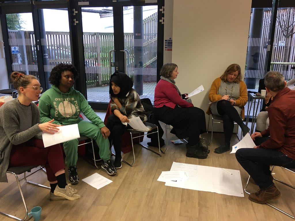 Six team members sat in a semi-circle in The Venue and working together. They hold sheets of paper and one large sheet is on the floor.