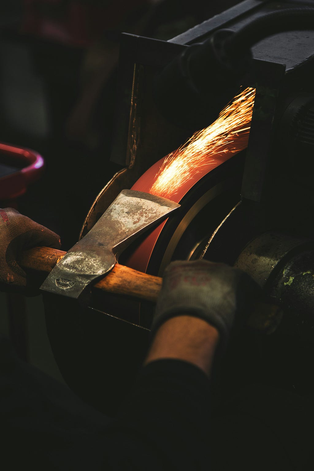 A craftsperson sharpens the blade of a small axe-head on a mechanical lathe, the grays of the image contrasted with the warm colors of the jet of sparks produced from the friction.