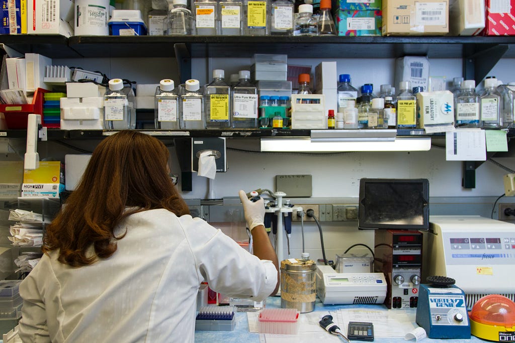 A woman standing at a lab table with shelves and shelves of bottles and other lab instruments, has her back to the camera and is dropping liquid into a glass beaker.