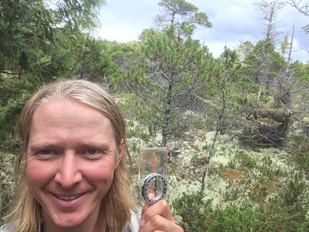 A selfie of a smiling person holding up a compass, with skinny trees and moss in the background.