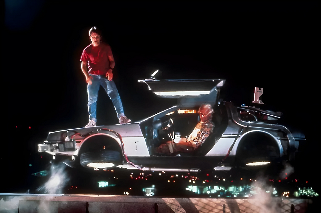 La imagen captura una escena de “Regreso al Futuro Parte II” donde Marty McFly está de pie sobre el capó del DeLorean, con las puertas abiertas. Dentro del coche, se encuentra Doc, preparado para ayudar a Marty en su huída.