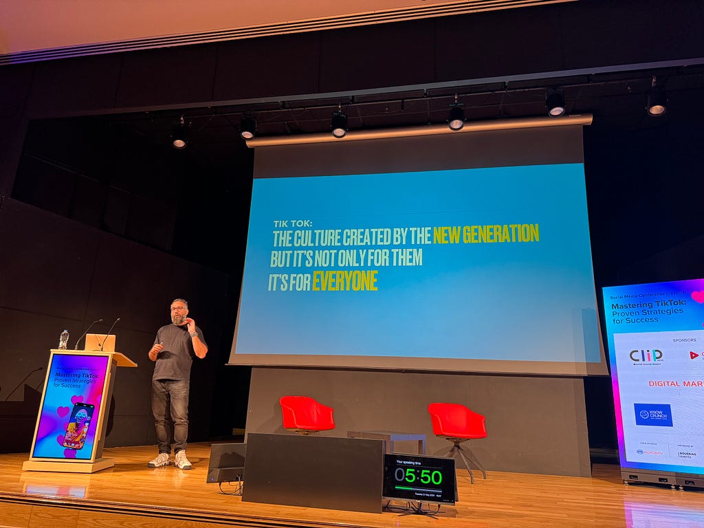 The image captures a speaker on stage who is standing to the left, gesturing with one hand, and a podium with a laptop is beside him. The slide on the large screen behind the speaker displays the following text: “TikTok: The culture created by the new generation but it’s not only for them. It’s for everyone.”