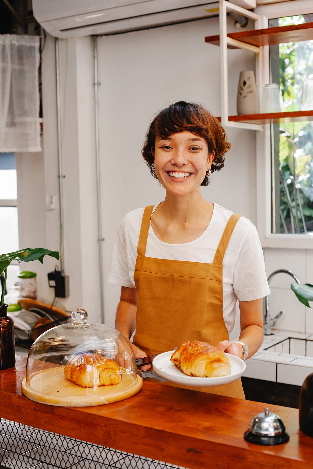 Smiling woman in an orange apron stands behind a wooden counter at what appears to be a bakery. Croissants line the counter. An open window is behind her to the right, a door to the left. White background with plants visible in the room.