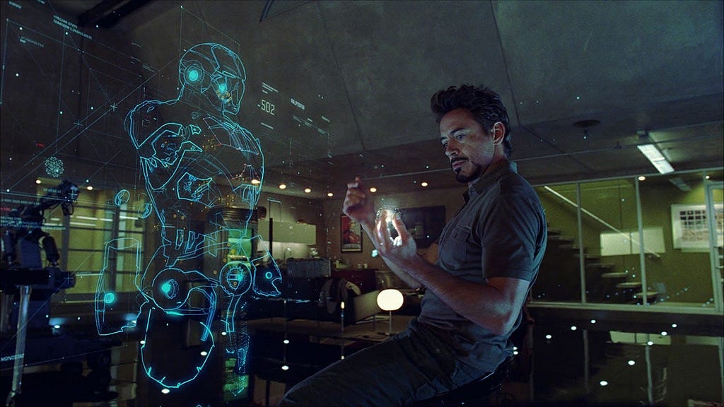 A still from Iron Man 2 showing J.A.R.V.I.S. (voiced by Paul Bettany) providing a translucent holographic iron-man suit interface on the workbench while Tony Stark looks on, illustrating how he seamlessly issues voice-commands to assemble and control different modules.
