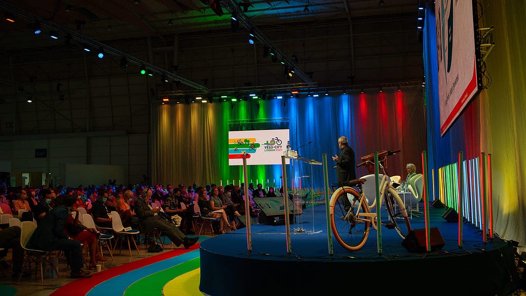 Conference room during the event Velo-city 2021 in Lisbon, with a bicycle on stage and colorful neon lights
