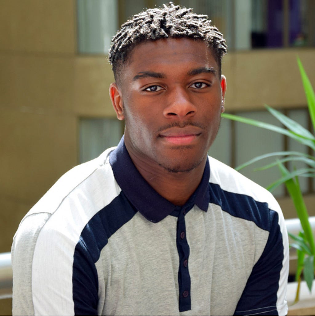 A portrait of Anthony, a young black man wearing a white collared shirt with navy trim.