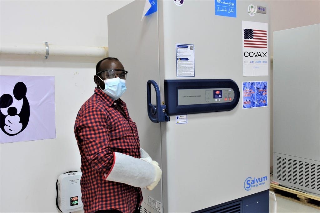 A man in goggles and mask wearing a large glove stands next to an ultra-cold chain freezer containing COVID-19 vaccines.