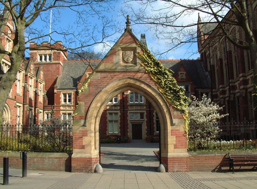 The archway of the Clothworkers South building stands on a sunny day. The brown and red brick is convered slighlty by ivy on the right side.