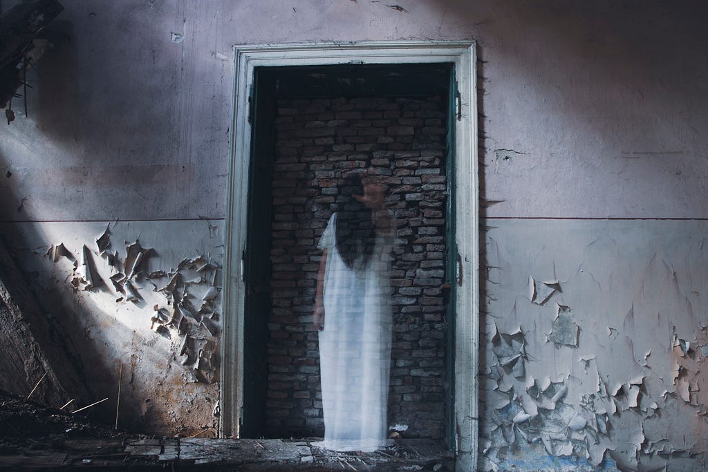 Image of a ghostly figure in a white dress standing in a doorway with a bricked-up entrance. The walls are peeling, adding to the eerie and abandoned atmosphere.