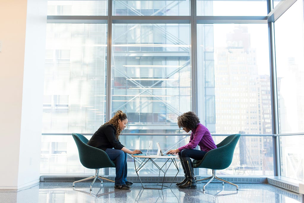 Two women sitting in green chairs next to floor to ceiling windows having a meeting.