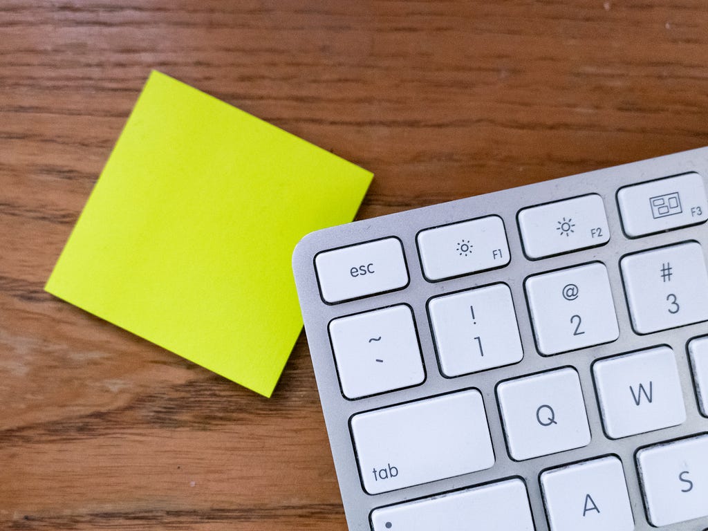 Photo of a computer keyboard with the tab and escape buttons in focus. The keyboard is overlapping a blank post-it note.