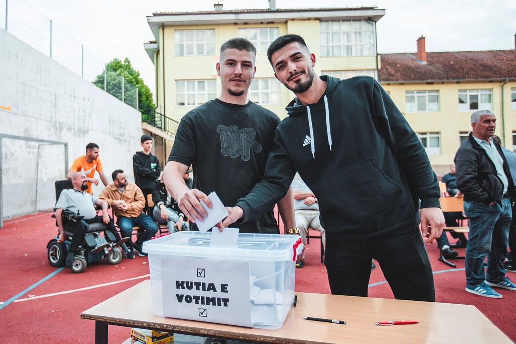 Two casually dressed men hold paper election ballots they are about to drop into a slot of a clear plastic box with a sign on it that reads “NUTIA E VOTIMIT,” Voting Notice.