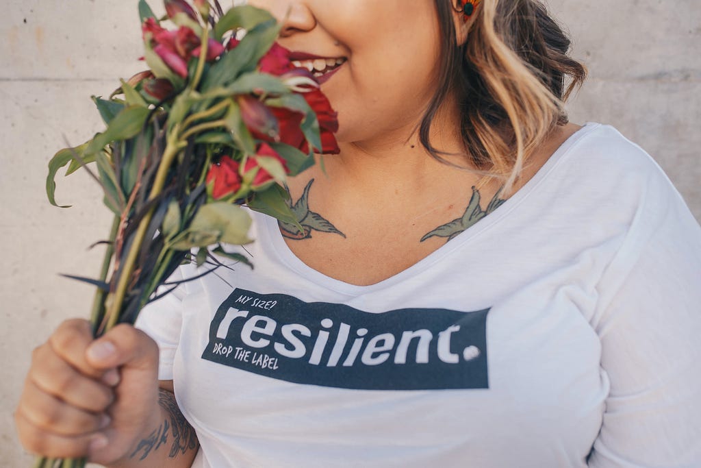 Uma mulher com camiseta branca escrito “resilient” cheira um buquê de rosas