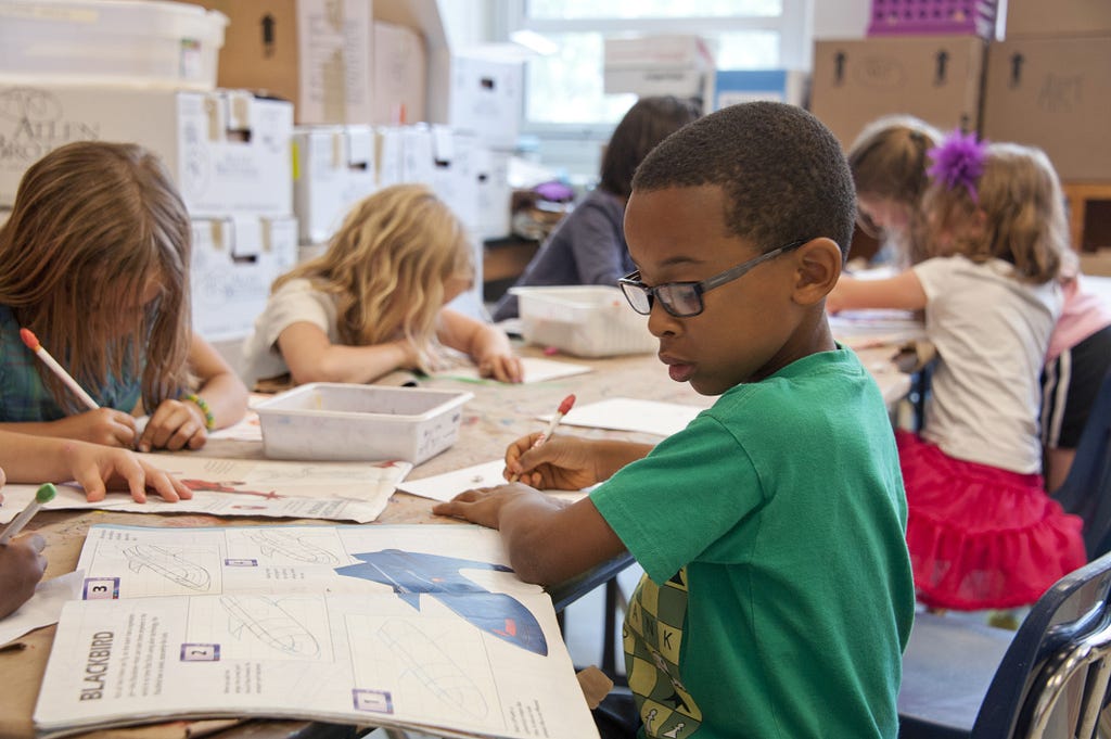 The picture shows 6 children working, independently, on some work. In the foreground is a young, black boy with a green shirt and glasses. He is focused on his work.