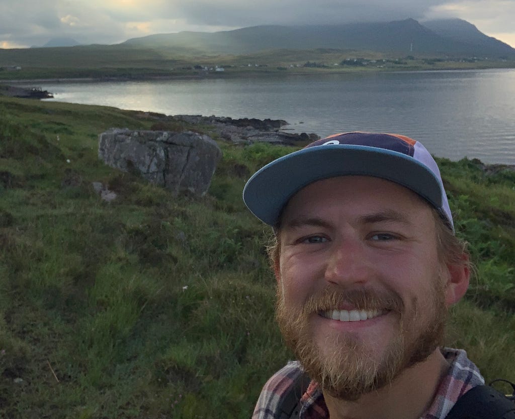 A hiker smiles at the camera beside a scottish sea loch, below brooding mountains.
