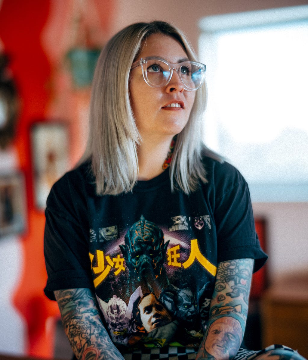 Erin sits with her hands folded in front of her, tattoos and vintage band shirt clearly visible, framed by orange and natural light.