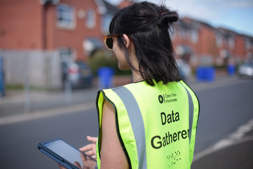 A person wearing a yellow high-visibility vest, printed with the words “Data Gatherer” and an Open Data Manchester logo, is standing on a residential street lined with red-brick houses, inputting data into a handheld electronic device.