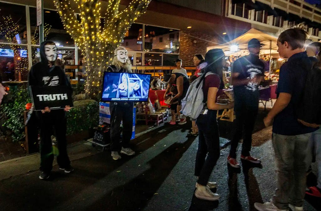 A photo of the Anonymous for the Voiceless, standing together. One on the left hand side holds a sign that reads “TRUTH”, while another holds a TV monitor showing an animal being slaughtered. They are standing next to a group of market-goers, who have their backs turned to them.