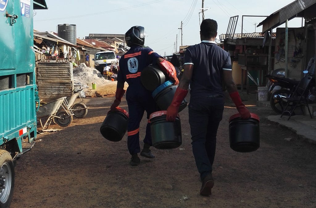 Two Clean Team employees carrying sanitation buckets down a road in Kumasi.