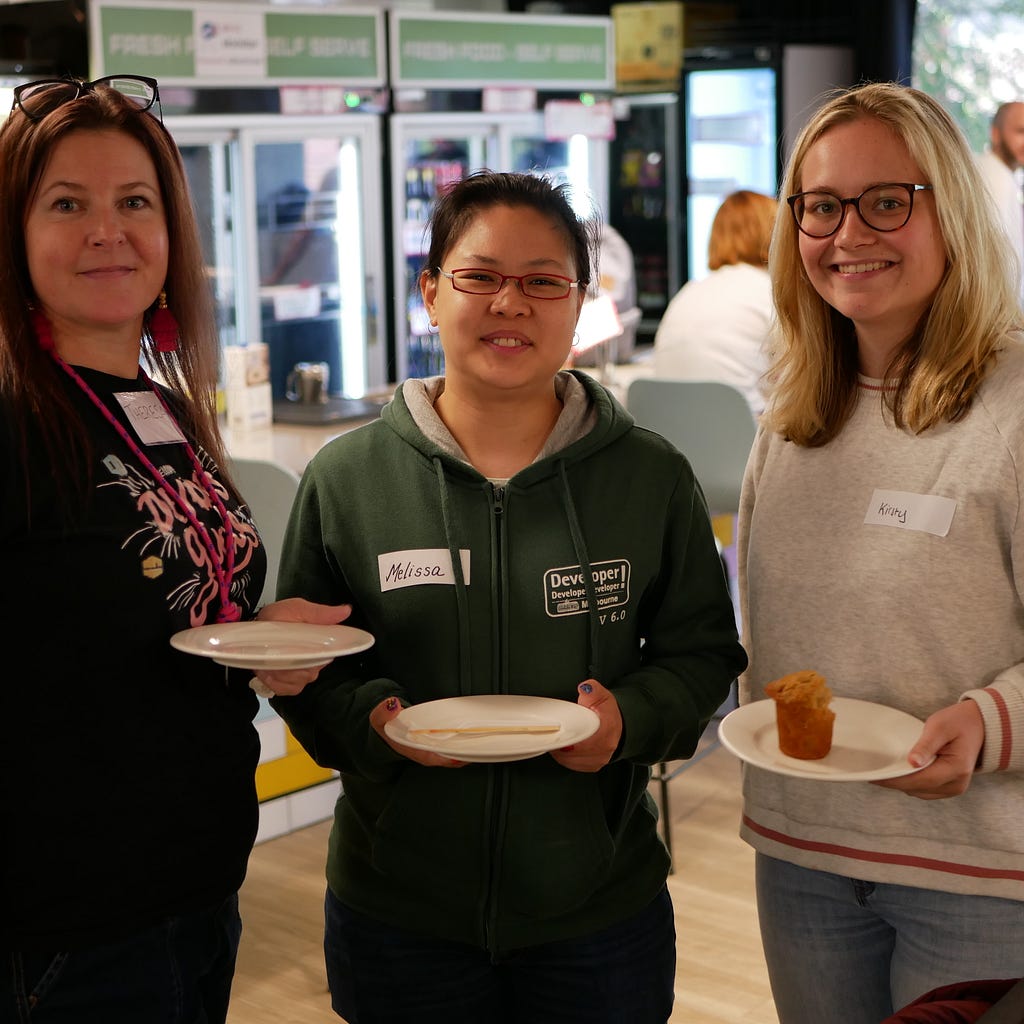 Three women are standing and holding plates with some food, smiling at the camera.