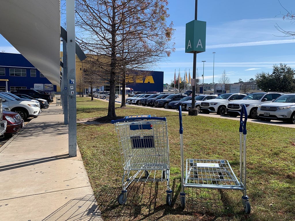Two carts sit abandoned on the grass median in front of a big box store parking lot. More carts are in the background.