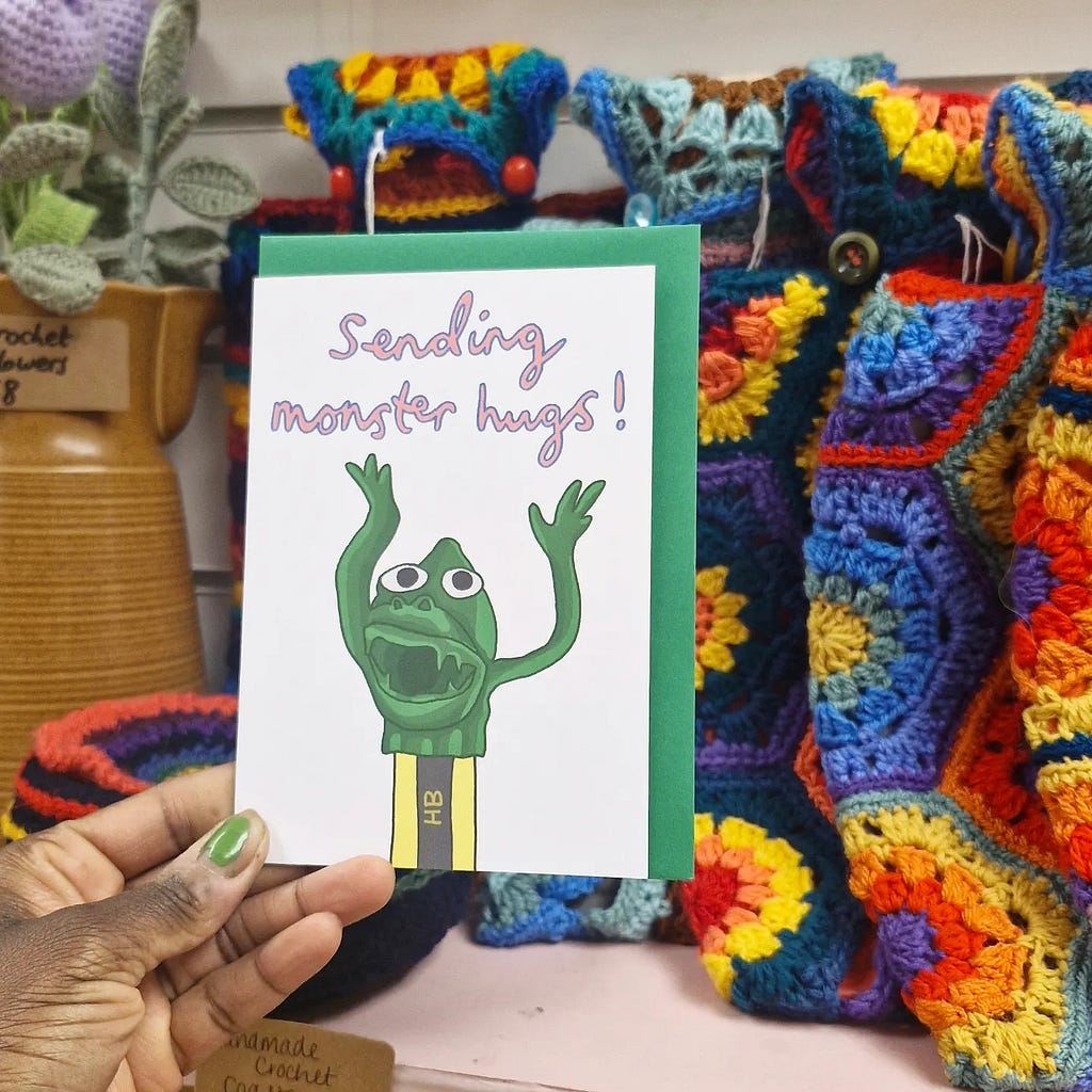 In the foreground, a white card with the message ‘sending monster hugs’. The card has a hand-drawn design of a novelty green monster, waving its long green arm on the top. The monster sits atop a pencil. There is a colourful crochet blanket in the background.
