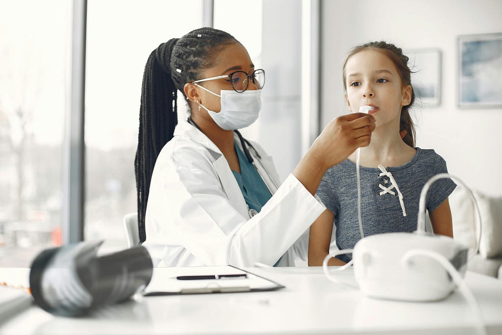 A physician wearing a masked gives a little girl an asthma test, she is blowing through a tube connected to a machine that measures the strength of her lungs.