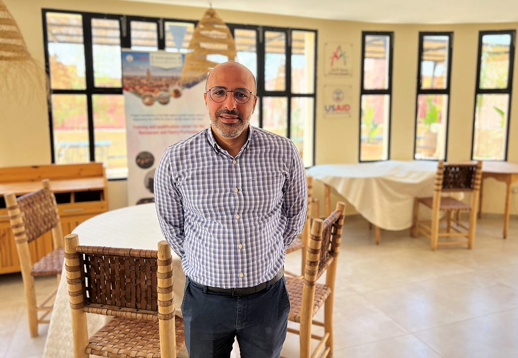 A Moroccan man stands in the center of an empty restaurant with the USAID logo in the background.